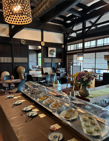 Una cafetería acogedora y tradicional con vigas de madera, una vitrina de pasteles, cerámica y flores. La luz natural entra por las ventanas, creando un ambiente cálido y acogedor con una decoración ecléctica.