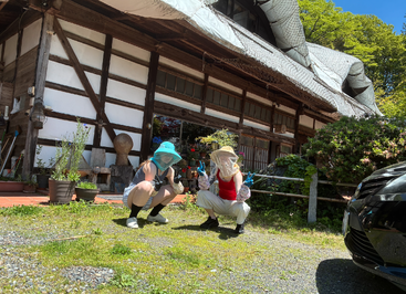 Dos personas están sentadas en cuclillas frente a una casa tradicional japonesa; llevan sombreros de ala ancha con red, guantes de jardinería y ropa informal. Están rodeadas de una exuberante vegetación y macetas.