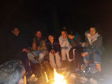 The image depicts a group of people gathered around a campfire at night, with some seated on logs and others standing nearby, all facing the fire.