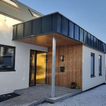 A modern house entrance with wooden paneling, a black balcony overhead, large windows, a concrete pathway, and two potted plants near the front door.
