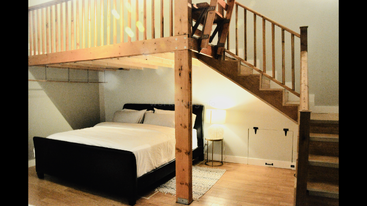 Esta acogedora habitación cuenta con una cama con marco negro, un altillo de madera con escaleras, una lámpara que proporciona una iluminación suave, suelos de madera noble, una decoración minimalista y paredes blancas impolutas.