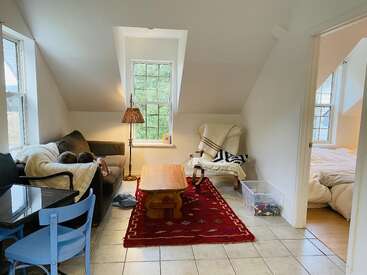 Un espace de vie confortable sous les combles comprend un canapé avec deux enfants, une table basse en bois, un tapis rouge, un fauteuil, un lampadaire, un ensemble de salle à manger et une chambre adjacente.