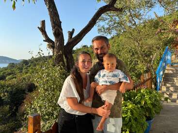 A happy family of three poses outdoors in bright sunlight, surrounded by lush greenery, trees, and blue skies, with a scenic hill and sea backdrop.