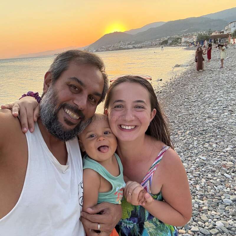 A joyful family of three smiles on a pebbled beach at sunset. Mountains and sea form the picturesque backdrop, radiating warmth, happiness, and togetherness.