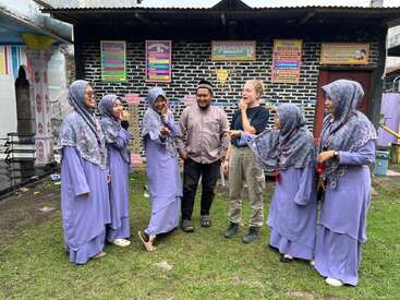 Eine Gruppe von Menschen, darunter Frauen in passenden lavendelfarbenen Kleidern und Kopftüchern, steht lachend und redend vor einem bunt geschmückten Gebäude.