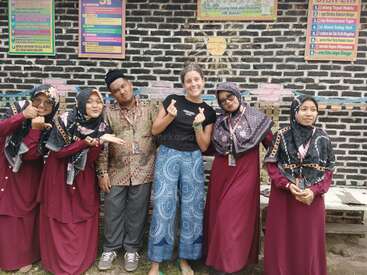 Eine Gruppe von sechs Personen posiert fröhlich vor einer Backsteinmauer mit bunten Schildern. Die meisten sind kastanienbraun gekleidet, einer trägt Freizeitkleidung.