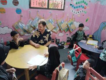Ein Mann sitzt mit kleinen Kindern an einem runden Tisch in einem farbenfrohen Klassenzimmer und beschäftigt sich mit ihnen. An der Wand hängen Kunstwerke, Bücher und Alphabetdekorationen.