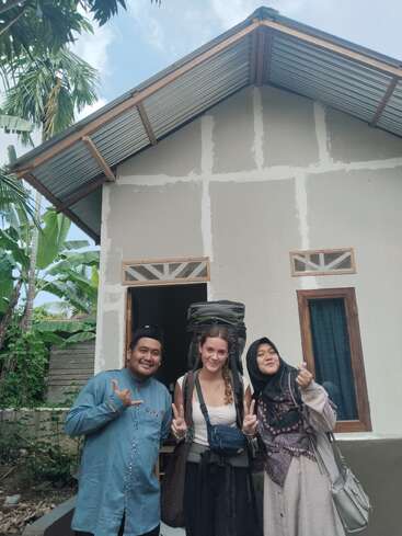 Three people pose happily in front of a house. The woman in the center has a backpack. The others make hand signs, and greenery surrounds them.