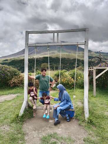 La imagen muestra a una familia de cuatro miembros posando en un columpio en un pintoresco entorno al aire libre, con un paisaje montañoso y un cielo nublado de fondo.