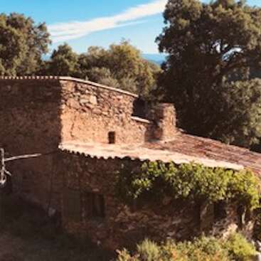Una rústica casa de piedra con tejado de tejas, rodeada de frondosos árboles y colinas, bajo la cálida luz del sol y un cielo azul despejado.