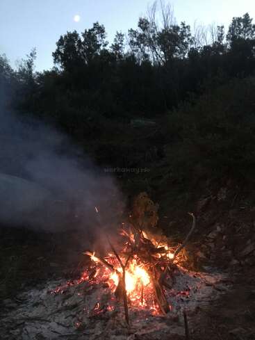 Una hoguera arde en el crepúsculo, rodeada de árboles y colinas. El humo se eleva hacia el cielo del atardecer, bajo el resplandor vigilante de la luna.