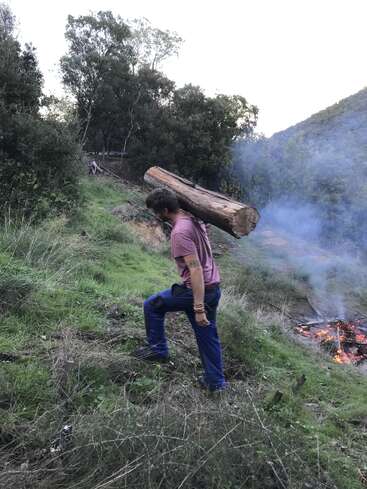 Un hombre lleva un gran tronco al hombro mientras camina cuesta arriba por una zona arbolada y cubierta de hierba, con una hoguera encendida y humo saliendo al fondo.