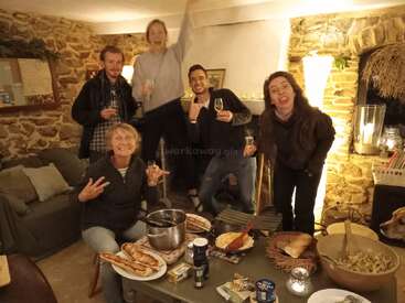 Cinco amigos celebrando en un acogedor salón rústico con paredes de piedra. Toman copas, sonríen y ponen caras tontas, rodeados de deliciosa comida y cálida iluminación.