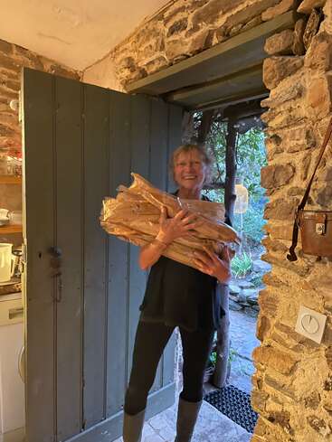 Una mujer sonriente se encuentra en un rústico portal de piedra, sosteniendo varias baguettes. Parece alegre, lleva ropa oscura, botas y está bañada por una cálida luz interior.