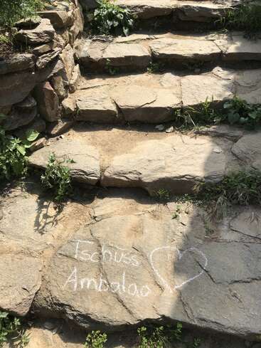 Esta imagen muestra unos escalones de piedra rodeados de vegetación. En uno de los escalones, "Tschüss Ambaloo" está escrito con tiza blanca junto a una forma de corazón.