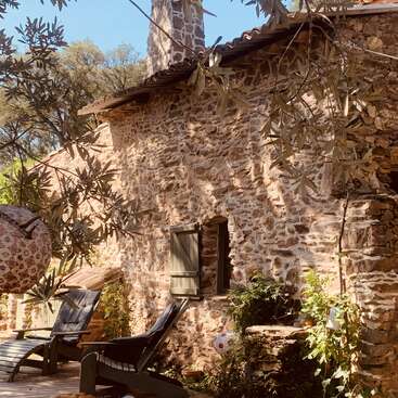 Cette image montre un chalet rustique en pierre avec une porte en bois, un patio ensoleillé, des chaises longues, des plantes feuillues et une lanterne décorative suspendue à un olivier.