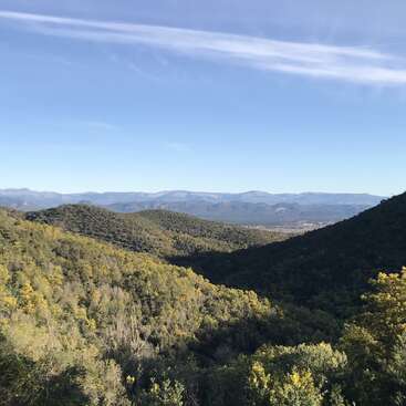 Esta imagem mostra uma paisagem cênica com colinas verdes cobertas de árvores, sob um céu azul claro com nuvens esparsas e montanhas distantes à vista.
