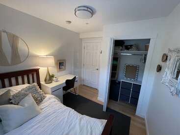 The image depicts a cozy bedroom with a bed, desk, and closet, featuring white walls, a dark wood headboard, and a black rug, exuding a sense of serenity and comfort.