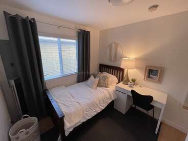 The image depicts a small bedroom with a bed, desk, and chair, featuring white walls, dark curtains, and a window, with a cozy and organized atmosphere.
