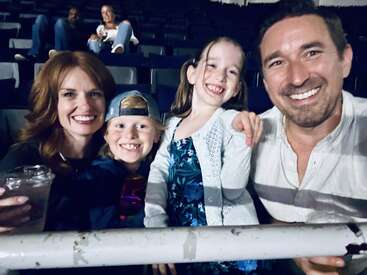 A happy family of four, smiling brightly, poses for a selfie at an indoor event. Two adults and two children enjoy their time together in the crowd.