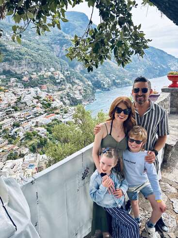 A smiling family of four poses together, wearing sunglasses, on a scenic balcony overlooking the colorful coastal town and beautiful mountains of Positano, Italy.