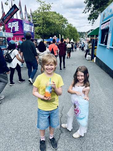 Two happy children stand at a busy fairground, smiling, holding a colorful slush drink and large blue cotton candy, surrounded by people and food stands.