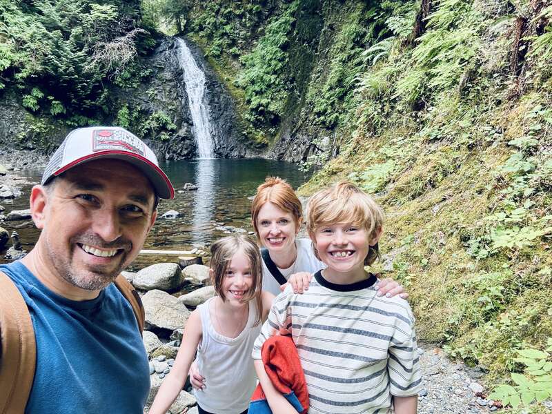 A smiling family of four poses in front of a scenic waterfall surrounded by lush greenery and rocks. They look happy, enjoying an outdoor adventure together.