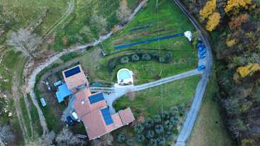 Esta imagen aérea muestra una casa con paneles solares, una piscina, coches aparcados, una carretera sinuosa, parcelas ajardinadas, hierba verde, árboles y bosques circundantes.