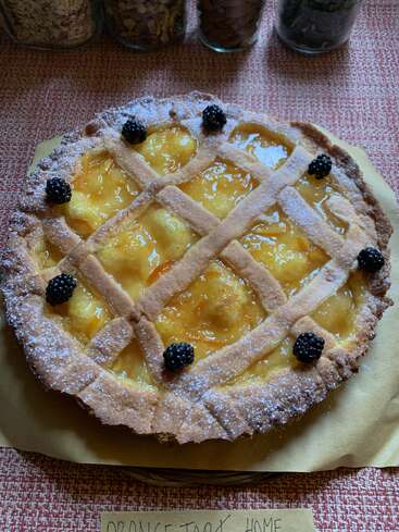 Das Bild zeigt eine selbstgemachte Orangentarte mit Gitterkruste, bestäubt mit Puderzucker und verziert mit frischen Brombeeren. Dahinter stehen Gläser mit Zutaten.