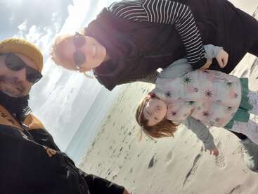 Una familia de tres personas sonríe en una playa de arena. Los adultos llevan gafas de sol y ropa de abrigo; el niño va bien abrigado y pone una cara juguetona. Brilla el sol.