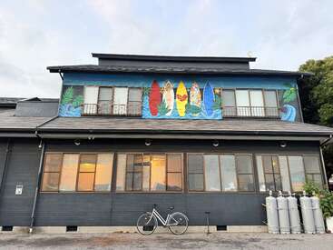 Esta imagen muestra un edificio con un mural de tablas de surf de colores en la fachada superior, ventanas, una bicicleta aparcada delante y varias bombonas de gas de gran tamaño.