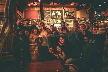 Un grupo de amigos posan juntos en un bar acogedor y poco iluminado, sonriendo y celebrando. Algunos sostienen una tarta, y un perro está presente.