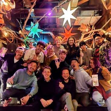 Un animado grupo de amigos posa en el interior bajo luces y adornos de colores en forma de estrella, sonriendo, riendo y haciendo gestos juguetones, capturando un momento alegre y festivo juntos.