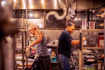 Dos chefs trabajan diligentemente en la bulliciosa cocina de un restaurante, rodeados de ollas, sartenes y utensilios, concentrados en preparar deliciosos platos en un ambiente cálidamente iluminado.