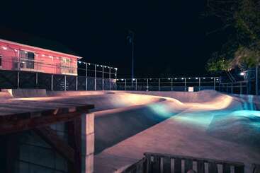 Un skatepark de hormigón liso se ilumina por la noche con luces de colores, que proyectan tonos azules, morados y cálidos. Un edificio rosa adyacente crea una atmósfera vibrante.
