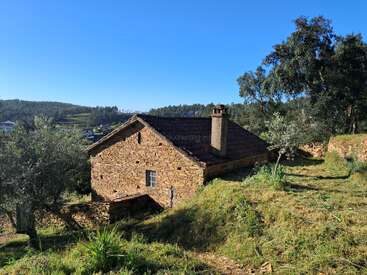 Ein rustikales Steinhaus steht auf einem grasbewachsenen Hügel, umgeben von Bäumen und üppiger Natur, unter einem strahlend blauen Himmel mit bewaldeten Hügeln in der Ferne.