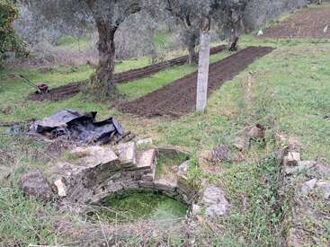 Das Bild zeigt einen rustikalen Garten mit umgegrabenen Beeten, einem alten Steinbrunnen mit grünem Wasser, Olivenbäumen, einem Rasenmäher und verstreuten schwarzen Planen in der Nähe.