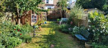 Um jardim exuberante com grama verde, flores coloridas, um galpão de madeira, uma árvore, equipamentos de recreação infantil, um banco verde e a luz do sol projetando sombras suaves.