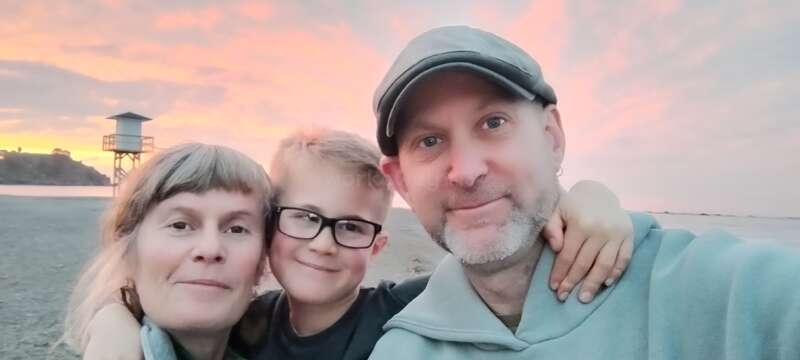 A smiling family of three poses for a selfie on a beach at sunset, with pastel skies and a lifeguard tower in the background, looking happy.