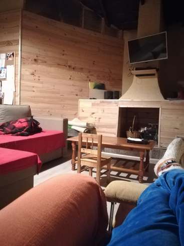 La imagen muestra una acogedora habitación con chimenea, televisor y dos camas, y en primer plano las piernas de una persona vestida con pantalones azules y calcetines peludos.