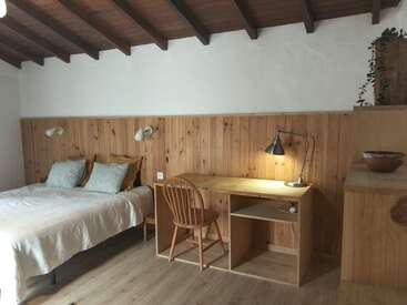 This cozy bedroom features a wooden desk with a lamp, a wooden chair, a comfortable bed with pillows, wood paneling, and a rustic, minimalist design.