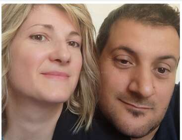 Un selfie en gros plan de deux personnes, une femme aux cheveux clairs et un homme aux cheveux foncés, qui regardent tous deux l'appareil photo avec un léger sourire.