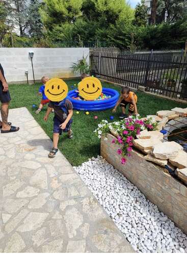 Des enfants jouent dans un jardin où se trouve une petite piscine bleue remplie de balles colorées. Les visages de deux enfants sont recouverts de smileys jaunes dessinés.