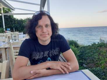 A man with curly hair and a nose ring sits at an outdoor restaurant overlooking the ocean, wearing a black band t-shirt and sunglasses on his head.