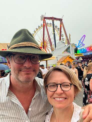 Ein lächelndes Paar steht auf einem lebhaften Jahrmarkt. Hinter ihnen bilden ein Riesenrad und bunte Lichter eine festliche Kulisse mit vielen Menschen, die das Ereignis genießen.