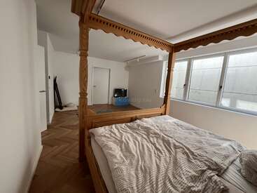 Dieses Bild zeigt ein gemütliches Schlafzimmer mit einem hölzernen Himmelbett, zerknittertem Bettzeug, großen Fenstern, einem Holzboden, einem kleinen Fernseher und einer minimalistischen Einrichtung, die eine friedliche Atmosphäre schafft.