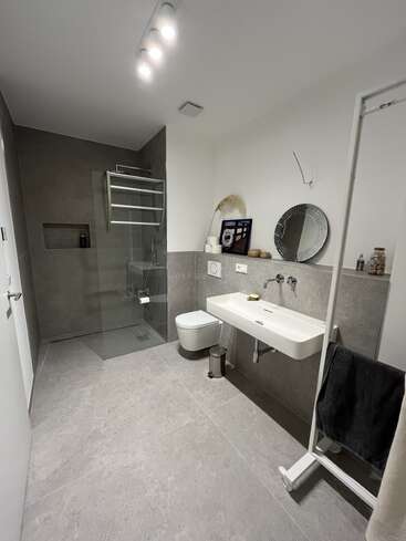 Dieses moderne Badezimmer hat große graue Fliesen, eine begehbare Glasdusche, eine wandmontierte Toilette, ein minimalistisches Waschbecken, einen runden Spiegel, Dekoartikel, einen Handtuchhalter und eine helle Deckenbeleuchtung.