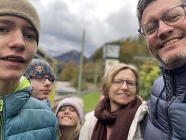 Eine fünfköpfige Familie lächelt an einem bewölkten Tag im Freien, warm gekleidet in Jacken und Mützen, zusammenstehend mit Bergen und einer Uhr im Hintergrund.