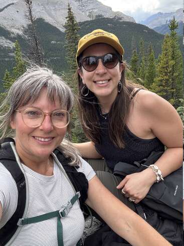Deux femmes posent joyeusement pour un selfie lors d'une randonnée en montagne, entourées d'arbres. Elles portent toutes deux un sac à dos ; l'une d'elles a des lunettes de soleil et un chapeau jaune.