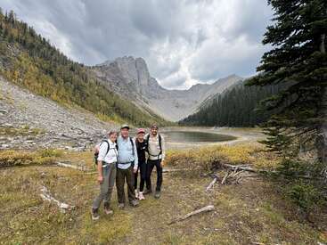 Quatre randonneurs posent en souriant devant un lac alpin pittoresque, entouré de montagnes escarpées, de pentes boisées et d'un ciel nuageux spectaculaire, capturant une belle aventure en plein air.